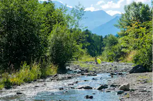 Ein Fluss mit Steinen und klarem Wasser fließt durch dichtes Grün, im Hintergrund Berge und bewaldete Hänge.