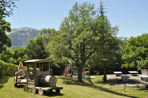 Grüner Campingplatz mit Rasen, Bäumen und Sitzgelegenheiten; im Vordergrund ein Holzunterstand und ein Grillwagen.
