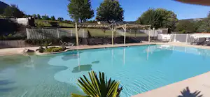 Schwimmbecken auf einem Campingplatz mit Palmenblatt im Vordergrund, Zaun und Liegen am Beckenrand, dahinter Wiesen und Bäume.