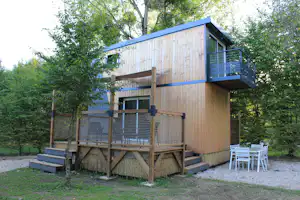 Holzhaus auf Stelzen mit Balkon und Terrasse, daneben Gartenmöbel, umgeben von Bäumen im Campingbereich.