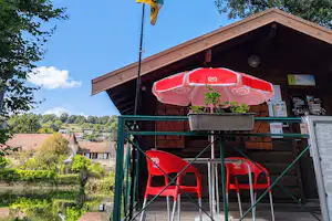 Camping-Rezeption mit rotem Sonnenschirm, Sitzgruppe und Infotafeln unter einem Holzvordach, im Hintergrund Bäume und Häuser.