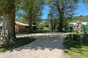 Campingplatz mit großen Bäumen, Kiesweg und Zeltstellplätzen, im Hintergrund kleine Gebäude und Schuppen.