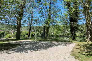 Schattiger Campingplatz mit Kiesweg und Bäumen, Blick auf Wiese und Landschaft im Hintergrund.