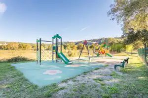 Spielplatz am Campingplatz mit Rutsche, Klettergerüst und Sandfläche auf gepflastertem Boden, umgeben von Wiese und Bäumen.