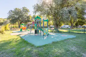 Spielplatz auf dem Campinggelände mit Rutsche und Klettergerüst, umgeben von Bäumen und Rasenfläche.