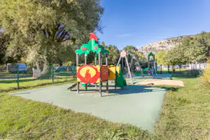 Spielplatz mit bunten Kletter- und Rutschfiguren auf dem Campingplatz, umgeben von Rasen, Bäumen und einem Zaun.