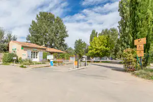 Camping-Eingang mit Schranke, Weg und Gebäude mit Ziegeldach, umgeben von Bäumen und Grünflächen.