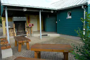 Überdachter Grillplatz mit gemauertem Kamin, Holztischen und Bänken auf gepflastertem Weg vor grüner Hütte.