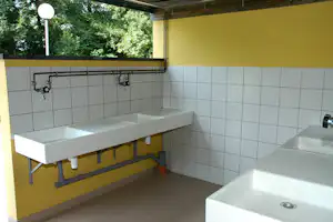 Waschraum mit mehreren Waschbecken, Armaturen und Fliesenwand unter einem gelben Dach, im Hintergrund Bäume.
