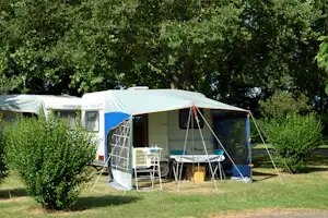 Camping-Caravan mit Vorzelt und Seitenwänden, davor Campingmöbel und Sonnenschirm im Grünen zwischen Büschen und Bäumen.