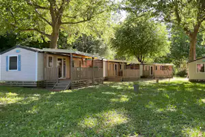Reihe von Mobilheimen mit Holzterrassen und Stufen auf einem grünen Campingplatz, umgeben von Bäumen und Rasen.