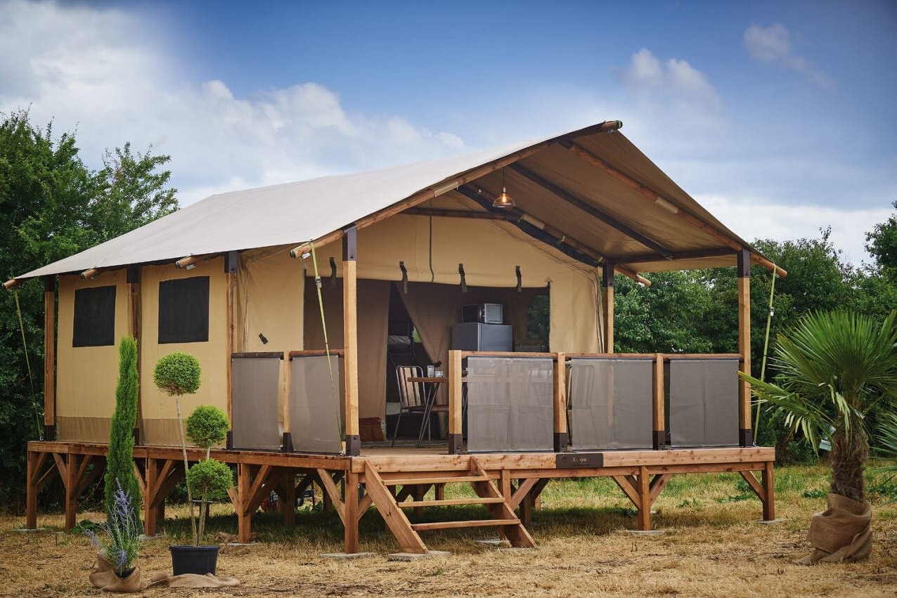 Zeltunterkunft auf Holzpodest mit Veranda, seitlichen Vorhängen und Sitzbereich; umgeben von Bäumen und Sträuchern im Campinggelände.