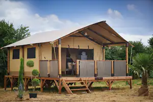 Zeltunterkunft auf Holzpodest mit Veranda, seitlichen Vorhängen und Sitzbereich; umgeben von Bäumen und Sträuchern im Campinggelände.
