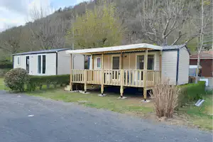Mobilheim mit Holzveranda und Sitzbank am Stellplatz, umgeben von Bäumen und Hügeln im Hintergrund.