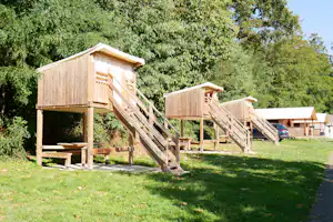 Holz-Spielhäuser mit Rutsche und Treppe auf einer Wiese, umgeben von Bäumen und einem Weg am Campingplatz.