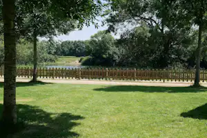 Grüne Campingwiese mit Holzlattenzaun, Bäumen im Vordergrund und Blick auf einen ruhigen Wasserlauf in der Ferne.