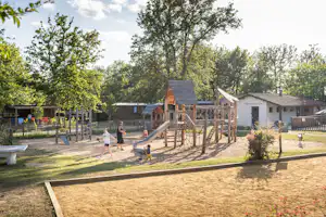 Spielplatz mit Klettergerüst und Rutsche, Sandboden, Bäume und ein Gebäude im Hintergrund auf dem Campinggelände.
