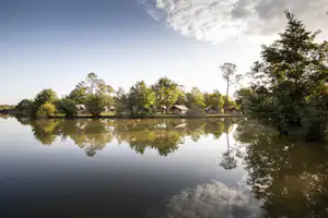 Ruhiger See mit Spiegelung, am Ufer kleine Hütten und Bäume, im Hintergrund weitere Gebäude am Campingplatz.