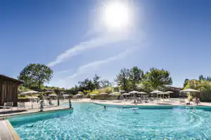 Schwimmbecken eines Campingplatzes mit Sonnenschirmen, Liegen und Bäumen im Hintergrund.