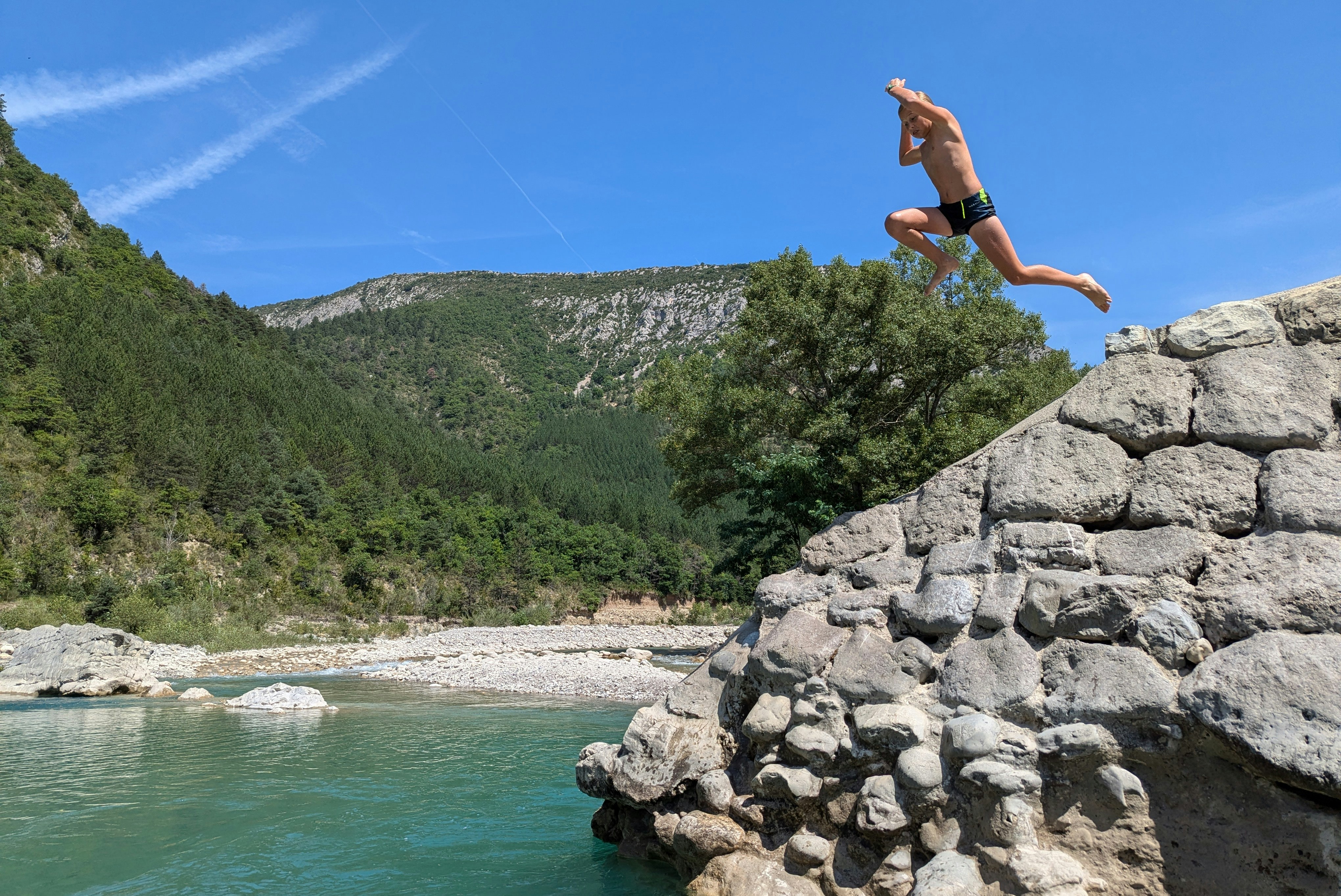 Ein Mann springt vom Felsen in den Fluss, umgeben von Bäumen, Bergen und klarem blauem Himmel.