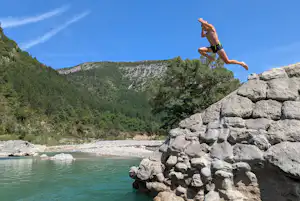Ein Mann springt vom Felsen in den Fluss, umgeben von Bäumen, Bergen und klarem blauem Himmel.