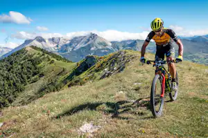 Radfahrer auf einem Bergpfad, grünes Gras und Berge im Hintergrund, weite Täler unter blauem Himmel.