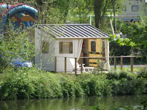 Kleines Camping-Häuschen mit Terrasse und Vorhängen am See, umgeben von Büschen und Bäumen im Garten.