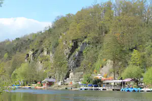 Campingplatz am See mit Steg, Booten und Bäumen am Ufer, im Hintergrund bewaldete Felswand und Hütten.