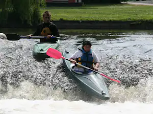Zwei Kinder paddeln mit Kajaks auf einem Fluss, im Hintergrund grünes Ufer und ein Campinggebäude.