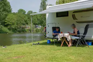 Wohnwagen am Flussufer im Camping, mit Tisch und Stühlen, zwei Personen sitzen draußen vor dem geöffneten Vorzelt.