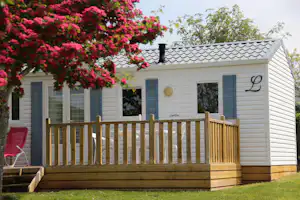 Mobilheim mit Holzterrasse, weißen Wänden und blauen Fensterläden, im Vordergrund rote Blüten und ein roter Liegestuhl.