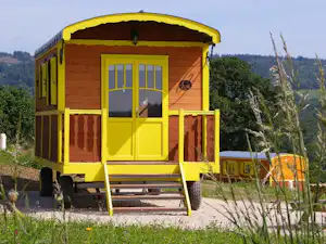 Gelber Wohnwagen mit Veranda und Eingangstür, davor Kiesweg und orangefarbene Markise, im Hintergrund Bäume und Hügel.