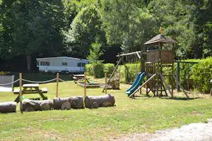 Spielplatz am Campingplatz mit Rutsche und Klettergerüst, umgeben von Bäumen und Rasen, im Hintergrund ein Wohnwagen.