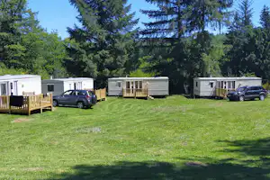 Mehrere Wohnwagen auf einem Campingplatz mit Holzterrassen, Rasenfläche und geparkten Autos vor dichtem Wald.