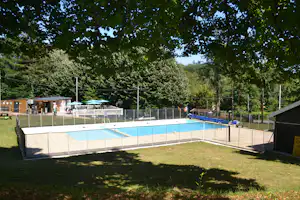 Schwimmbecken auf dem Campingplatz mit Umzäunung, Liegen und Sonnenschirmen, umgeben von Bäumen und Rasen.