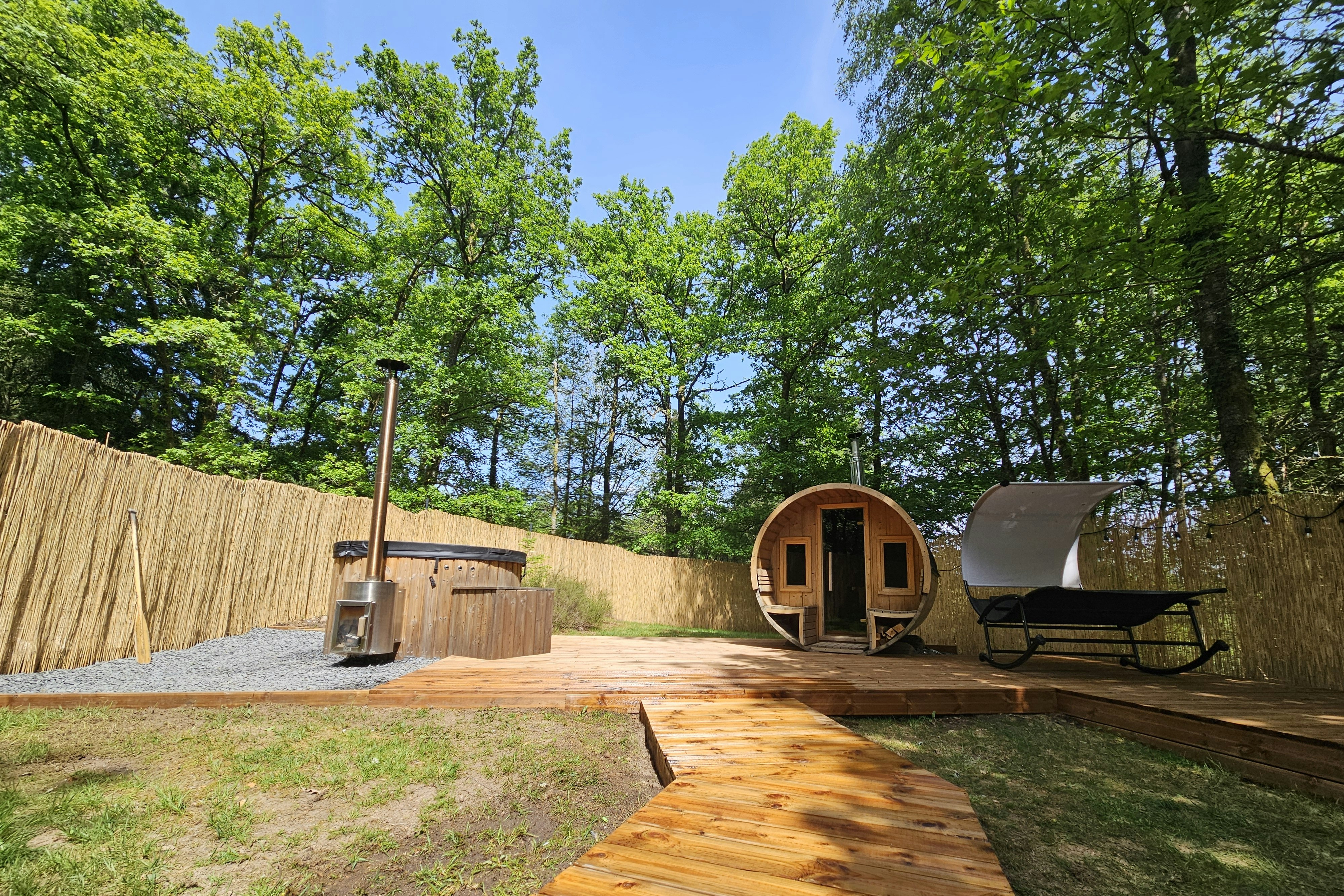 Holzterrasse mit Campingmöbeln, davor ein Zeltanhänger und ein Sonnenschirm, umgeben von Hecken und Bäumen im Garten.