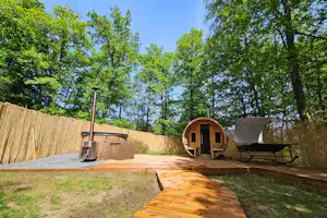 Holzterrasse mit Campingmöbeln, davor ein Zeltanhänger und ein Sonnenschirm, umgeben von Hecken und Bäumen im Garten.