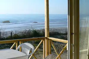 Blick von der Camping-Terrasse aufs Meer, mit Holzkonstruktion, Tisch und Stuhl vor dem Strand und Felsen im Wasser.