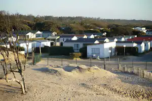 Reihen weißer und blauer Ferienhäuser auf einem Campingplatz, umzäunte Wege und Sandfläche mit Bäumen im Vordergrund.