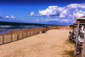 Strandpromenade am Meer mit Holzzaun, Sandweg und Blick auf die Küste, daneben ein Fahrradständer.