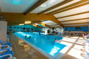 Hallenbad mit blauem Schwimmbecken, Liegen und Sitzbereichen unter Holzdecken, mehrere Personen im Wasser.