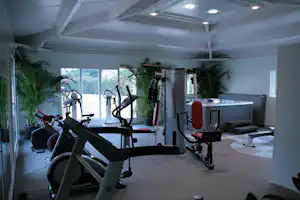 Fitnessraum im Campingbereich mit Fahrradergometern, Kraftgeräten und großen Fenstern, daneben eine Bank und Hanteln.