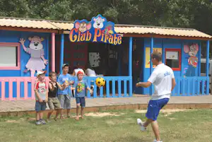 Kinder spielen vor einem bunten Spielhaus mit Piratenmotiv, blauer Umrandung und Sitzbank auf dem Campingplatz.