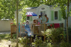 Wohnwagen mit Terrasse im Grünen, davor Holzgeländer und Pflanzen, im Hintergrund zwei Personen am Eingang.