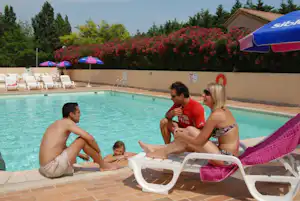 Familie am Camping-Pool auf Liegen, im Hintergrund Sonnenschirme und Hecken, daneben ein Gebäude mit Holzfassade.