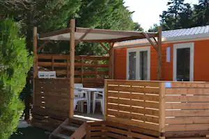 Holzbungalow mit Terrasse und Holzgeländer, davor Stühle und Tisch, umgeben von Bäumen und Hecken am Campingplatz.