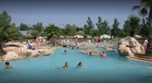 Großes Camping-Schwimmbecken mit Felsen und Palmen, Gäste im Wasser, daneben Liegen und Gebäude im Hintergrund.