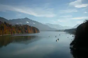 Ruhiger See mit kleinen Booten, Uferbäumen in Herbstfarben und Bergpanorama im Hintergrund.