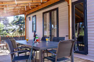 Terrasse am Campinghaus mit Tisch, Stühlen und Glastüren, im Hintergrund Bäume und Holzverkleidung.
