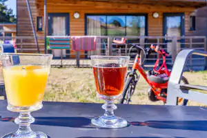 Zwei Gläser mit Getränken auf einer Camping-Terrasse, im Hintergrund Holzhaus und Gartenstühle.